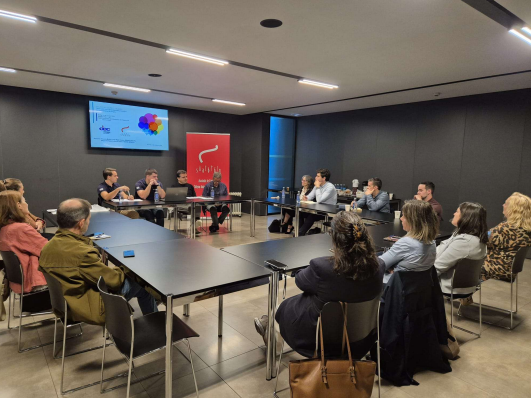 Na imaxe vese unha reunión de traballo nunha sala de reunións, na que varias persoas están sentadas arredor dunha mesa disposta en forma de U. As persoas asistentes escoitan as intervencións e participan no intercambio de ideas. Na parte frontal da sala hai unha pantalla con información dunha presentación e, ao seu carón, un expositor institucional. O espazo, amplo e ben iluminado, favorece o diálogo, a participación e o traballo en equipo.